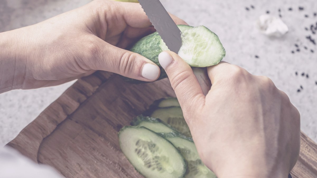 Manos cortando un pepino sobre una tabla de madera, con limón, ajo y verduras alrededor
